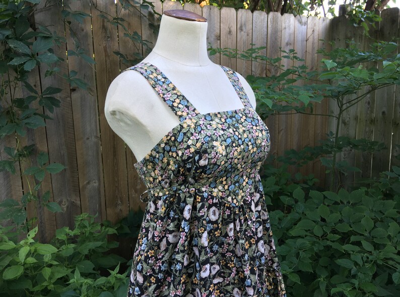 Puede incluir: Vestido de verano con estampado floral sobre un maniqu&iacute;. El vestido presenta un fondo negro con un estampado de flores blancas, azules y amarillas. Tiene un escote cuadrado, tirantes anchos y cintura fruncida. El vestido se exhibe al aire libre.