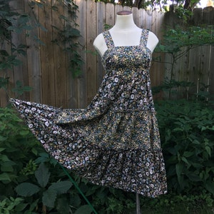 Puede incluir: Vestido midi con estampado floral, escote cuadrado y tirantes anchos. El vestido presenta una falda escalonada y se exhibe en un maniqu&iacute;. La tela tiene un fondo negro con peque&ntilde;as flores amarillas, blancas y azules.