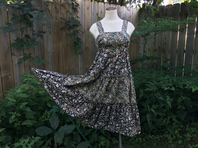 Puede incluir: Vestido midi con estampado floral, escote cuadrado y tirantes anchos. El vestido presenta una falda escalonada y se exhibe en un maniqu&iacute;. La tela tiene un fondo negro con peque&ntilde;as flores amarillas, blancas y azules.