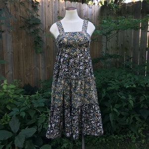 Puede incluir: Vestido con estampado floral sobre un maniqu&iacute;. El vestido presenta un fondo negro con un estampado de flores blancas, amarillas y azules. Tiene tirantes anchos y una falda escalonada, creando una silueta fluida. El vestido se exhibe al aire libre contra una valla de madera y vegetaci&oacute;n.