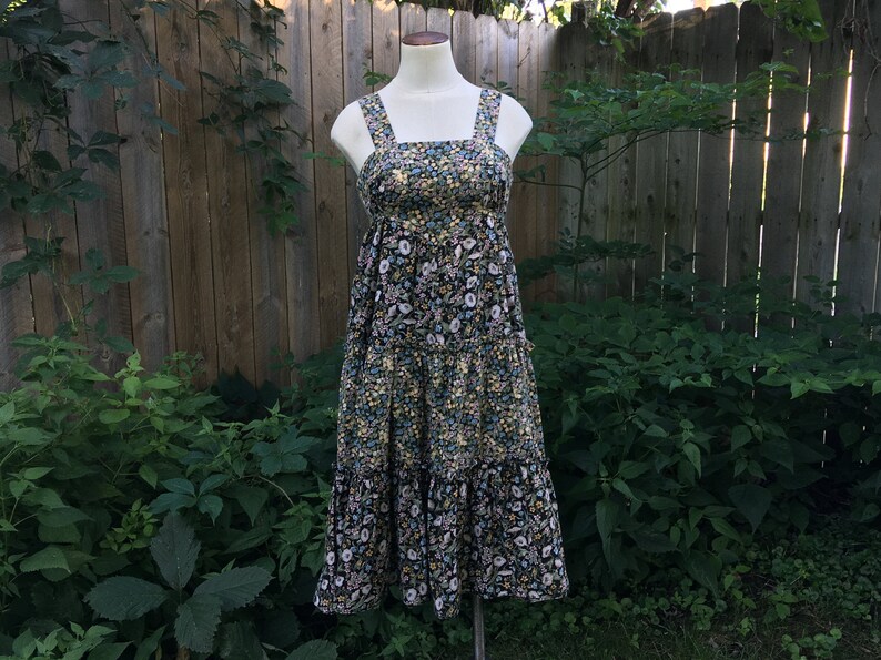 Puede incluir: Vestido con estampado floral sobre un maniqu&iacute;. El vestido presenta un fondo negro con un estampado de flores blancas, amarillas y azules. Tiene tirantes anchos y una falda escalonada, creando una silueta fluida. El vestido se exhibe al aire libre contra una valla de madera y vegetaci&oacute;n.