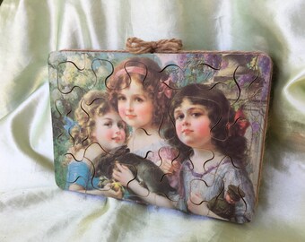 May include: A wooden jigsaw puzzle with a vintage illustration of three girls. The girls are wearing dresses and have long hair. The puzzle is made up of many pieces and is in a frame.