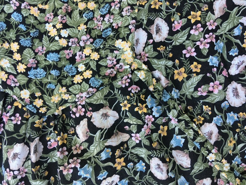 Puede incluir: Primer plano de una tela con un estampado floral. El dise&ntilde;o presenta un fondo negro con una variedad de flores en tonos blancos, rosas, amarillos y azules, junto con hojas verdes. La tela parece ser un material ligero.