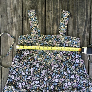 Puede incluir: Vestido con estampado floral sobre fondo negro, con peque&ntilde;as flores azules, amarillas y rosas. El vestido tiene tirantes anchos y un dobladillo con volantes. Una cinta m&eacute;trica indica un ancho de unos 38 cm.