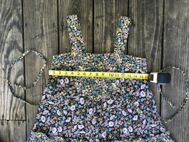 Puede incluir: Vestido con estampado floral sobre fondo negro, con peque&ntilde;as flores azules, amarillas y rosas. El vestido tiene tirantes anchos y un dobladillo con volantes. Una cinta m&eacute;trica indica un ancho de unos 38 cm.
