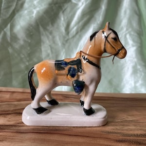 May include: A ceramic horse figurine with a tan and white body, black mane and tail, and a blue and black saddle. The horse has a brown bridle and is standing on a white rectangular base. The figurine is a decorative item.