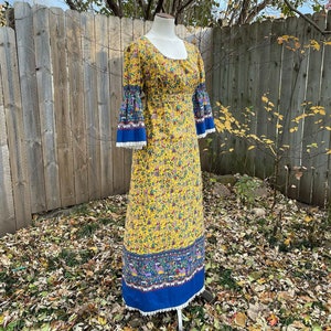 May include: A yellow maxi dress with a floral print and blue trim. The dress has bell sleeves and a gathered waist.