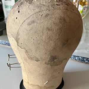 May include: A vintage wig head form, featuring a light tan fabric surface with dark stains and a black base. The head form has metal pins and a stitched seam around the crown. The form is designed for wig styling and display.