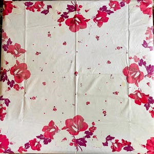 May include: White tablecloth with a floral pattern featuring large red and pink flowers and smaller red dots. The flowers are clustered in the corners and along the edges of the cloth, with purple leaves.