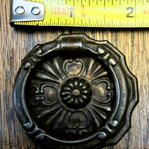 May include: Antique bronze-colored cabinet pull with a floral design. The pull is round with a central flower and radiating petals. A yellow tape measure is above the pull, showing 1 inch.