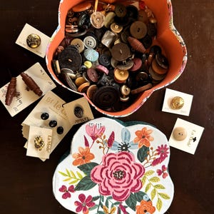 May include: A flower-shaped orange container filled with various buttons in shades of brown, black, and beige. Several buttons are displayed on cards, and a floral-decorated container is also present. The buttons vary in size and material.