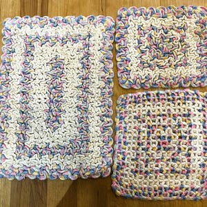 May include: Four handmade crochet washcloths in a variety of patterns. The washcloths are primarily off-white with accents of pink, blue, and yellow. Each cloth has a scalloped edge in coordinating colors. The textures vary from tight to open weave.