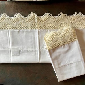 May include: White pillowcases with a cream-colored, crocheted lace trim. The set includes two pillowcases, one folded and one unfolded, showcasing the intricate lace design. The pillowcases are made of a smooth, white fabric.