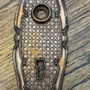 May include: An elongated, metallic door plate with a dark bronze finish, featuring an intricate lattice pattern across the center, a circular knob opening at the top, and a keyhole opening at the bottom, accented by ornate scrollwork at both ends.