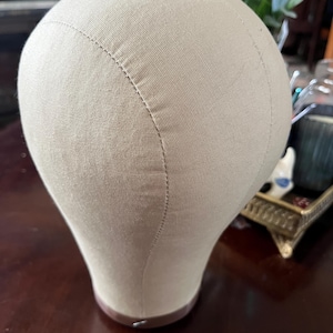 May include: A beige canvas wig head form, designed to resemble a human head. The form has a rounded top and a neck, with visible stitching along the seams. The base is a darker brown color.