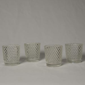 May include: Four clear glass votive candle holders. Each holder has a textured, bubble-like pattern on the exterior. The holders are cylindrical with a slightly flared top and sit on a flat base. They are arranged in a row against a white background.