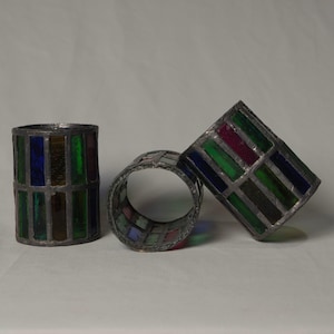 以下が含まれることがあります： 青、緑、赤、黄色のステンドグラスのナプキンリングが3つ。リングは金属フレーム付きの円筒形で、1つは直立、1つは平ら、1つは傾いています。