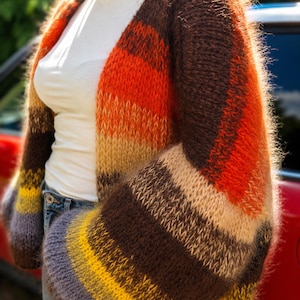 May include: A fuzzy, open-front knit cardigan featuring horizontal stripes in shades of dark brown, bright orange, beige, yellow, and grey. The garment has a relaxed fit with wide sleeves and a soft, textured yarn construction worn over a white shirt.