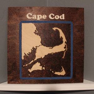 May include: A wooden sign with a burnt brown background and a light brown outline of Cape Cod. The sign has a blue border and the words "Cape Cod" are written in white.