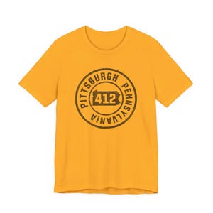 May include: A mustard yellow short-sleeve t-shirt featuring a circular dark brown graphic on the chest. The design includes the text "PITTSBURGH PENNSYLVANIA" surrounding a silhouette of the state of Pennsylvania with the number "412" inside.