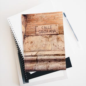 May include: A notebook with a cover depicting a weathered brick wall and a sign reading "CALLE SANTA ANA." The notebook is stacked with others, and a pen lies on the white surface beside it. The cover has a spiral binding.