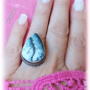 Puede incluir: Un anillo de plata con una piedra de ágata blanca y gris en forma de lágrima engastada en un engaste de plata. El anillo está en una mano con esmalte de uñas blanco.