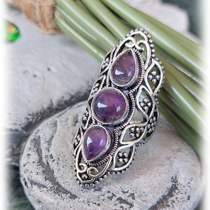Puede incluir: Un anillo de plata con un diseño intrincado que presenta tres piedras de amatista. El anillo está colocado sobre un fondo de piedra gris.