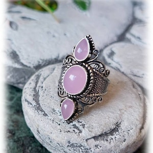 Puede incluir: Un anillo de plata con tres piedras rosas. El anillo tiene un diseño delicado y ornamentado.