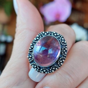 Puede incluir: Anillo ovalado con una gran gema rosa iridiscente. El anillo tiene un engaste detallado de color plateado con un borde decorativo. La palabra "BARQUENIKE" está impresa en la parte inferior de la imagen.