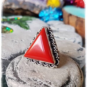 Puede incluir: Un anillo de plata con una piedra preciosa roja triangular engastada en un engaste de plata decorativo. El anillo está sobre un fondo de piedra gris.
