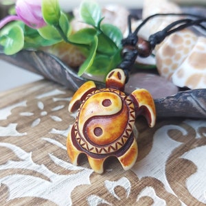 May include: A brown and white turtle pendant necklace with a yin yang symbol on its shell. The necklace is made of a black cord and is hanging on a wooden surface.