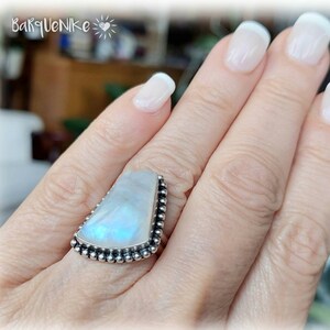 Puede incluir: Un anillo de plata con una gran piedra de luna iridiscente engastada en un engaste de cuentas. La piedra tiene forma triangular con una punta puntiaguda.