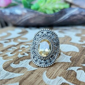 May include: A silver ring featuring an oval-shaped yellow gemstone set within an ornate silver bezel. The ring boasts a detailed design with small beads and swirls surrounding the gemstone.