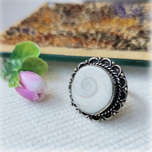 Puede incluir: Un anillo de plata con una incrustación de concha blanca. La concha tiene un patrón en espiral. El anillo tiene un borde decorativo.
