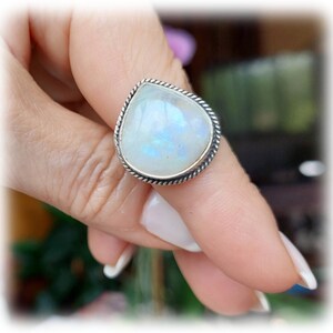 Puede incluir: Un anillo de plata con una piedra de luna en forma de lágrima. La piedra es iridiscente y tiene un color blanco, azul y gris.
