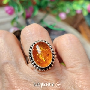 Puede incluir: Un anillo de plata con una piedra ovalada de ámbar engastada en un engaste de cuentas. La piedra de ámbar tiene un color cálido, similar al de la miel, y es translúcida.