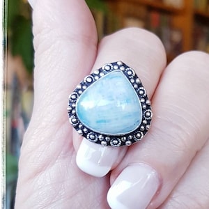 May include: A silver ring with a light blue gemstone in a triangular setting. The ring is on a finger with a white French manicure.