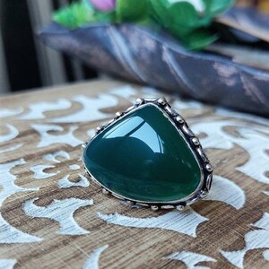 Puede incluir: Un anillo de plata con una piedra preciosa verde oscuro engastada en forma triangular. El anillo tiene un borde decorativo con pequeñas cuentas.