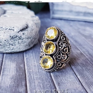 Puede incluir: Un anillo de plata con tres piedras preciosas amarillas engastadas en un diseño floral. El anillo está sobre una superficie de madera.
