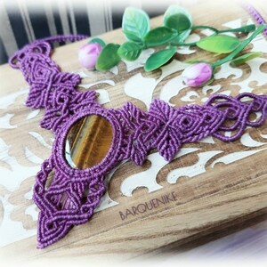 Peut inclure: Collier en macramé violet avec un pendentif en œil de tigre. Le collier est posé sur une surface en bois avec un motif floral blanc.