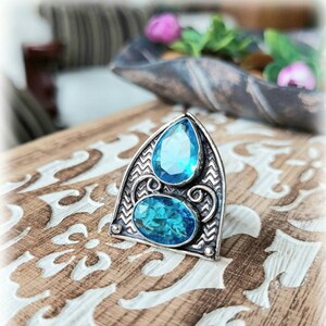 May include: A silver ring featuring a blue gemstone design. The ring has a pointed arch shape with intricate details and two blue gemstones.