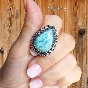 May include: A silver ring featuring a teardrop-shaped blue and green gemstone set within an ornate silver bezel. The ring is worn on a finger.