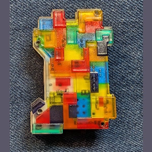 May include: A rectangular pin featuring a translucent, multi-colored block puzzle design. The accessory is composed of various interlocking geometric shapes in vibrant shades of red, blue, yellow, green, and orange, creating a mosaic effect.