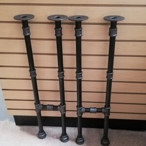 May include: Four black metal pipe legs with flanges on top. The legs are connected with pipe fittings to form a table base.