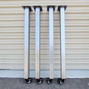 May include: Four vertical silver metal table legs with a square profile, each featuring a flat mounting plate at the top with four pre-drilled holes and a black swivel caster wheel with a white roller at the bottom for mobility and support.