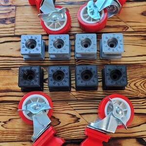 May include: An assortment of casters and mounting blocks. The casters have red wheels and brackets with silver hardware. There are also several gray and black mounting blocks. The items are arranged on a wooden surface.
