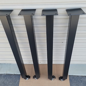 May include: Four black metal table legs with a square mounting plate and a tapered rectangular design. Each leg has a small wheel at the bottom. The legs are made of a matte black metal.