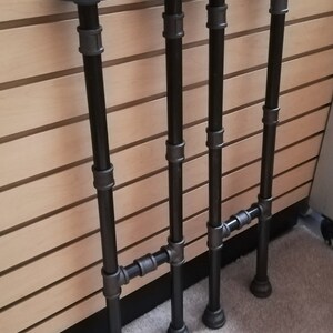 May include: Four black metal pipe table legs with flanges. Each leg is made of three sections of pipe connected by black metal fittings. The legs are arranged in a row, with the flanges facing the viewer.