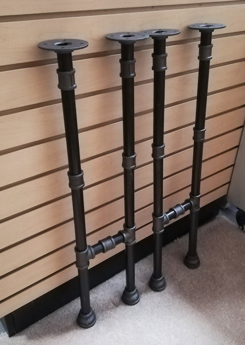 May include: Four black metal pipe table legs with flanges. Each leg is made of three sections of pipe connected by black metal fittings. The legs are arranged in a row, with the flanges facing the viewer.