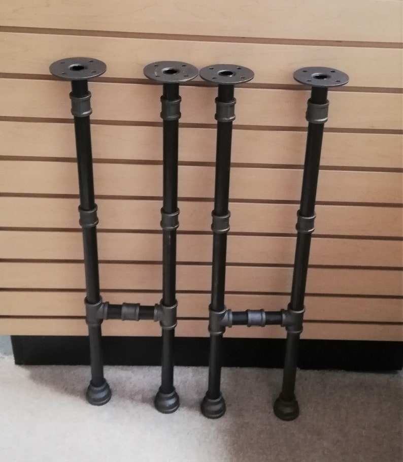 May include: Four black metal pipe table legs with a flange on top and a threaded end on the bottom. The legs are arranged in a pair of two, with the bottom pipes connected to form a cross.